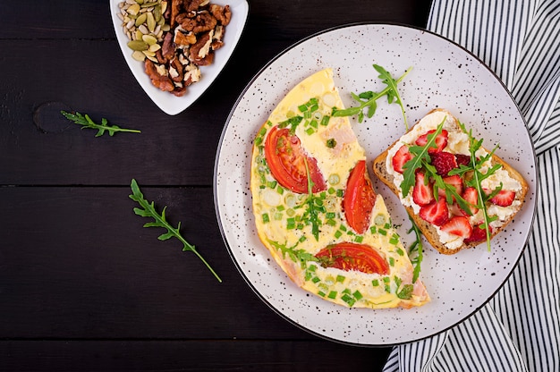 Omelet met tomaten, ham, groene ui en sandwich met aardbei op donkere tafel