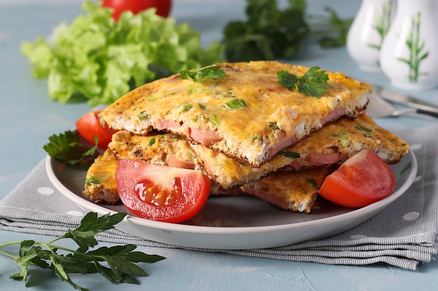 Omelet met worstjes, kaas en peterselie geserveerd met tomaten op een grijze plaat op een lichtblauwe achtergrond. Detailopname.