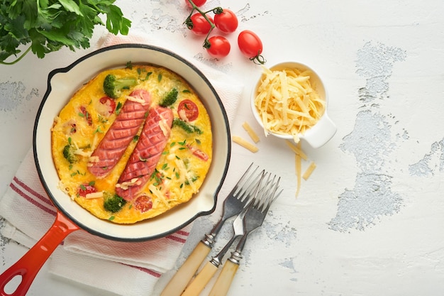 Omelet of frittata met twee worstjes gesmolten kaas kerstomaatjes broccoli en peterselie op rode koekenpan of ijzeren pan op witte oude betonnen achtergrond Gezond ontbijt eten concept Bovenaanzicht