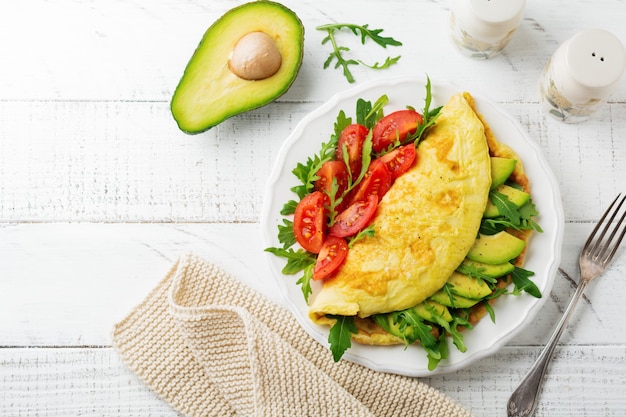 Omelette with avocado, tomatoes and arugula on white ceramic plate on light stone background