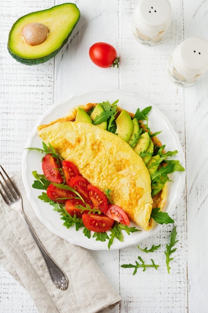 Omelette with avocado, tomatoes and arugula on white ceramic plate on light stone surface. Healthy breakfast