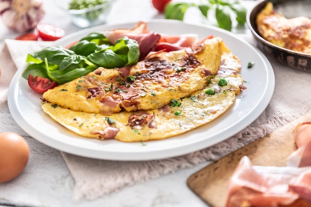 Omelette with prosciutto peas basil tomatoes and herbs on white plate.