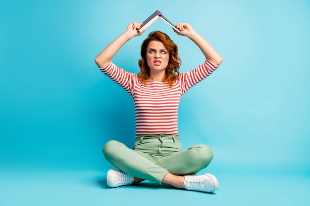 Omg examenvoorbereiding weer! Foto van het hele lichaam van boos vrouw zitten gekruiste benen kijken boven het hoofd houden papieren boek afkeer van haar encyclopedie dragen groene stijlvolle outfit geïsoleerde blauwe kleur