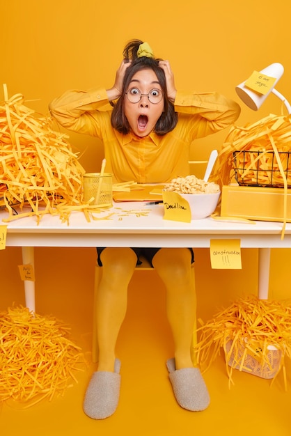 Omg how much work. Emotional scared woman looks with panic grabs head has deadline surrunded by sticky notes and cut paper poses at desktop against yellow background. Shocked female office worker