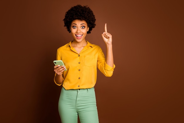 Omg ik weet welke tekst! Opgewonden gek afro Amerikaans meisje blogger gebruik smartphone type sms denk krijgen ongelooflijk idee vinger opsteken dragen gele stijlvolle groene outfit geïsoleerde bruine kleur