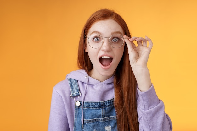 かっこいい。肖像画驚いた言葉のない興奮した赤毛の女の子ドロップジョー面白がって凝視カメラ驚いた印象的な投稿、オレンジ色の背景を読んで素晴らしい製品ネットタッチメガネを見つけます。