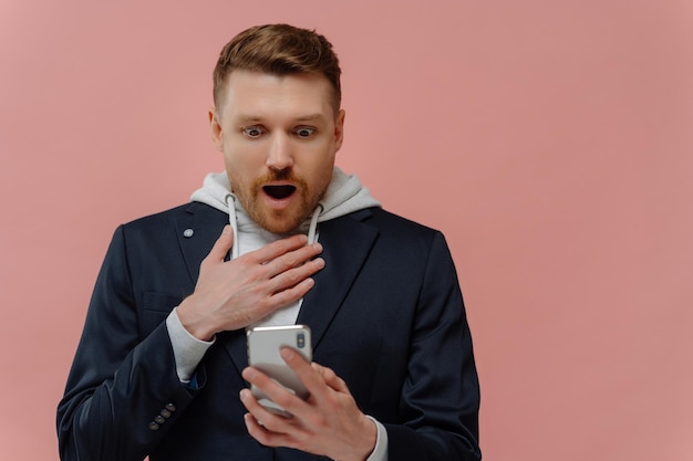 Omg what is it startled bearded european man cant understand
something on mobile phone stares at display cannot believe in
shocking news dressed in black jacket isolated over pink
background