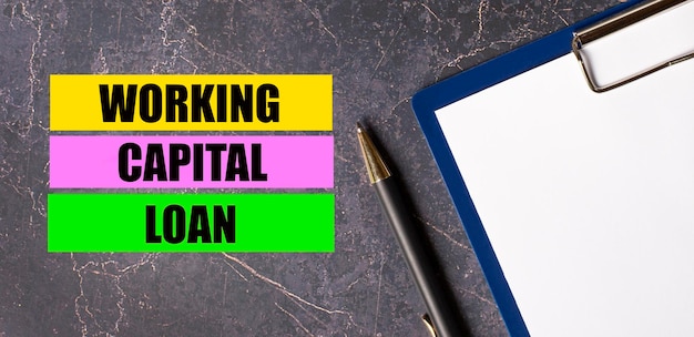 写真 暗い大理石の背景に、紙のペンが付いたタブレットと、「working capital loan」というテキストが記載された 3 つの明るい色とりどりの紙片