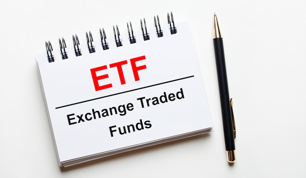 밝은 배경에 흰색 노트북에는 Etf Exchange Traded Funds라는 단어와 펜이 있습니다.
