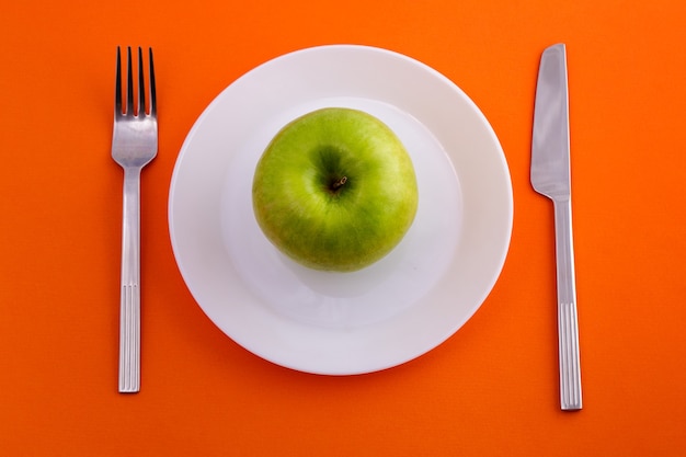 하얀 접시에 녹색 사과가 놓여 있고 상단 디에서 오렌지 배경 보기에 포크가 달린 칼이 있습니다.