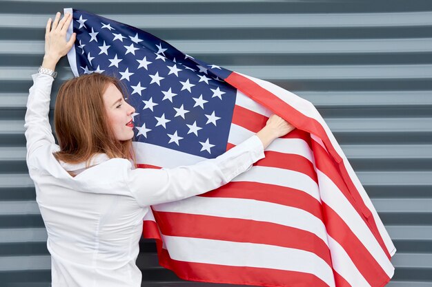 Onafhankelijkheidsdag en patriottisch concept. Jonge roodharigevrouw met rode geschilderde lippen die zich met gegolfd door de vlag van de windv.s. Bevinden