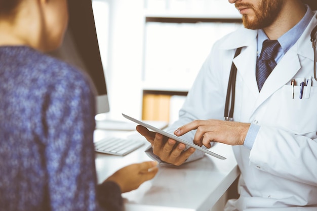 Onbekende arts met rode baard en geduldige vrouw bespreken het huidige gezondheidsonderzoek terwijl ze zitten en tabletcomputer gebruiken in de kliniek, close-up. geneeskunde concept.