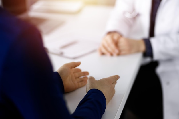 Onbekende vrouw-dokter praat met haar patiënt over haar diagnose, zittend aan het bureau in de zonnige kast in een kliniek. Arts op werkplek, close-up. Perfecte medische service i