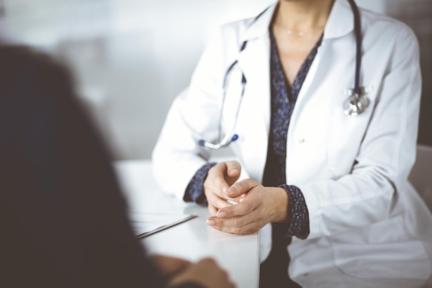 Onbekende vrouw-dokter praat met haar patiënt over haar diagnose, zittend samen aan het bureau in de kast in een kliniek. arts op de werkplek, close-up. perfecte medische service in een ho