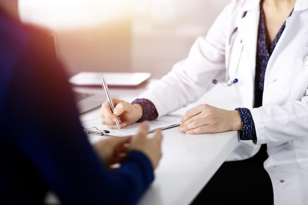 Onbekende vrouw-dokter schrijft enkele medische aanbevelingen aan haar patiënt, terwijl ze samen aan het bureau in de zonnige kast in een kliniek zitten. Arts gebruikt een klembord, close-up. Per