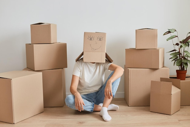 Onbekende vrouw met wit t-shirt zittend op de vloer in de buurt van kartonnen dozen, poserend met kartonnen doos op haar hoofd met tekenend gezicht erop, verhuizend naar een nieuw appartement.