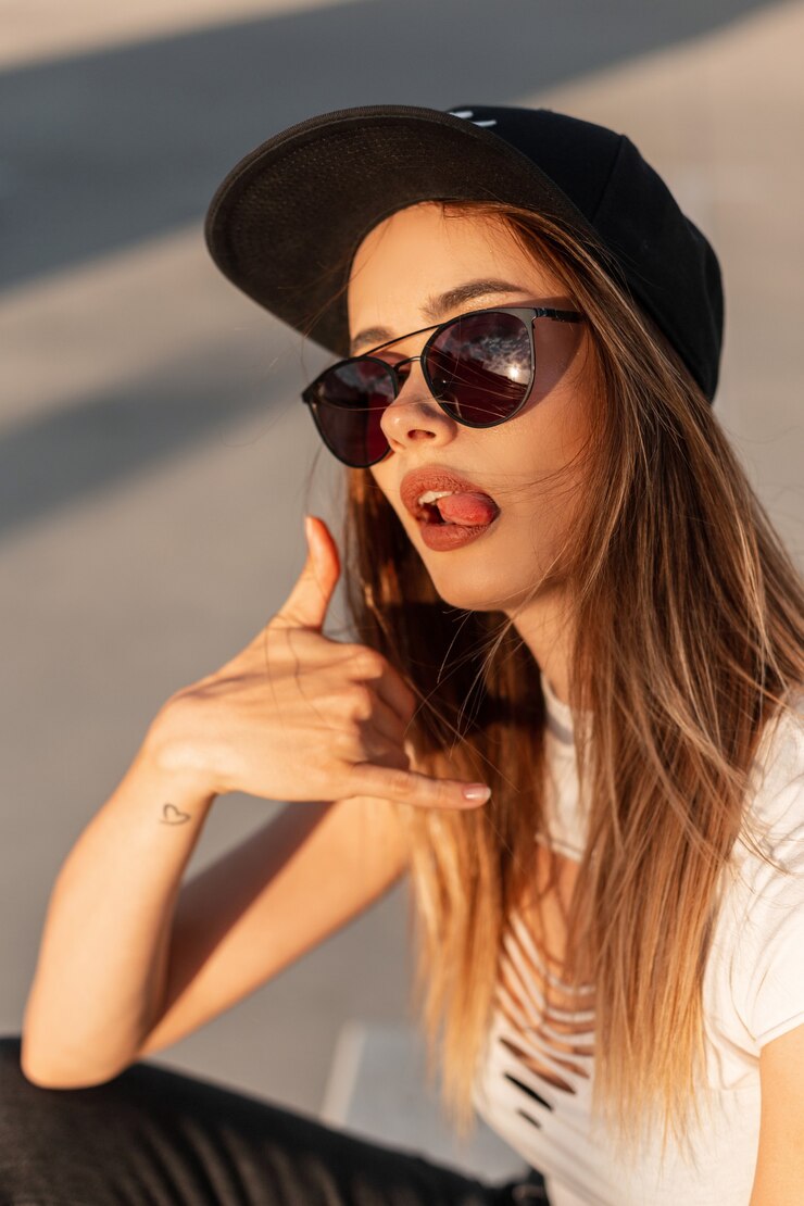 Foto onbeschaamd cool jong meisje hipster in modieuze pet in zonnebril in stijlvolle jeugd gescheurde top toont seksueel tong en toont "bel me". portret moderne vrouw bij zonsondergang.