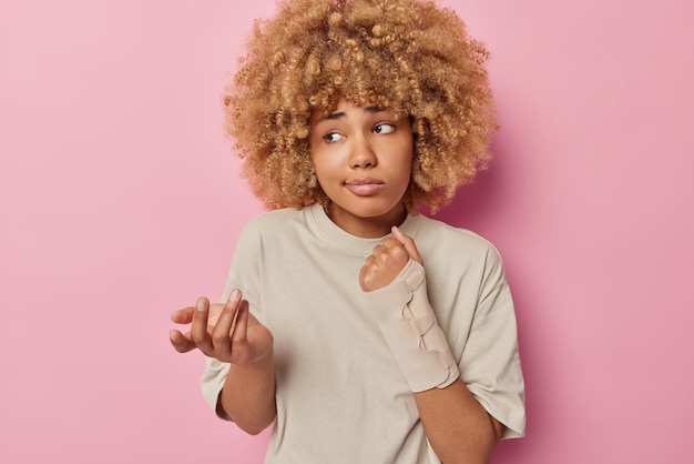 Foto onbewust krullend haired vrouw draagt elastische bandage op arm kreeg trauma na ongeval haalt schouders op, gekleed in casual t-shirt geïsoleerd over roze achtergrond letsel en gezondheidsproblemen concept