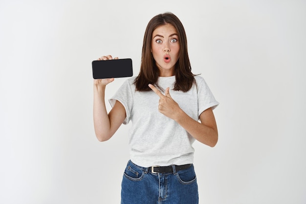 Onder de indruk brunette vrouw die het scherm van de mobiele telefoon toont en naar de mobiele telefoon wijst en verbaasd kijkt naar de witte achtergrond van de camera