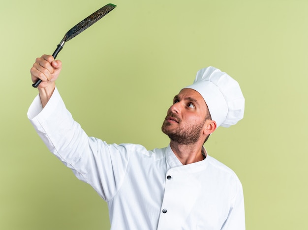 Onder de indruk jonge blanke mannelijke kok in chef-kok uniform en pet verhogen en kijken naar koekenpan