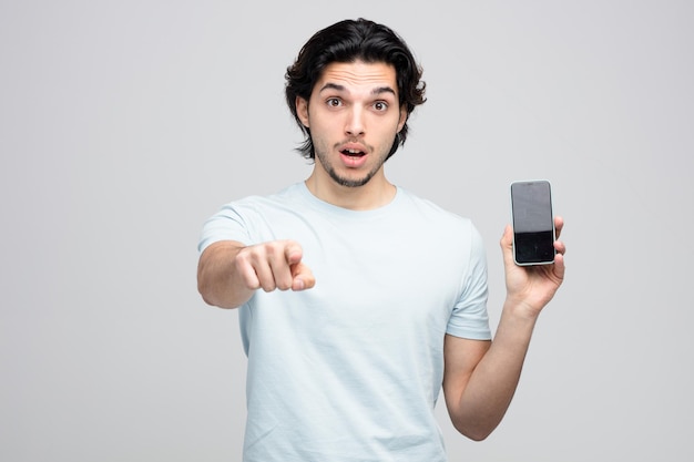 onder de indruk jonge knappe man met mobiele telefoon kijken en wijzend op camera geïsoleerd op een witte achtergrond