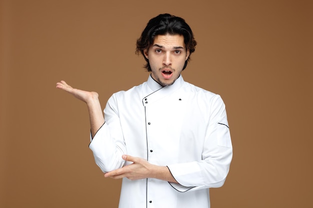 onder de indruk jonge mannelijke chef-kok in uniform kijkend naar camera met lege hand die elleboog aanraakt geïsoleerd op bruine achtergrond