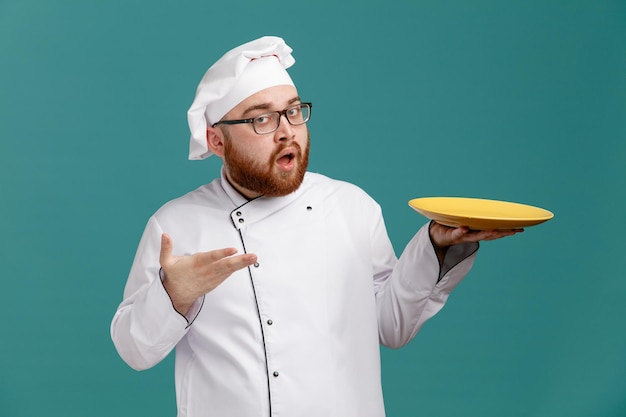 Onder de indruk jonge mannelijke chef-kok met een uniforme bril en een pet met een lege plaat die ernaar wijst met de hand kijkend naar camera geïsoleerd op blauwe achtergrond