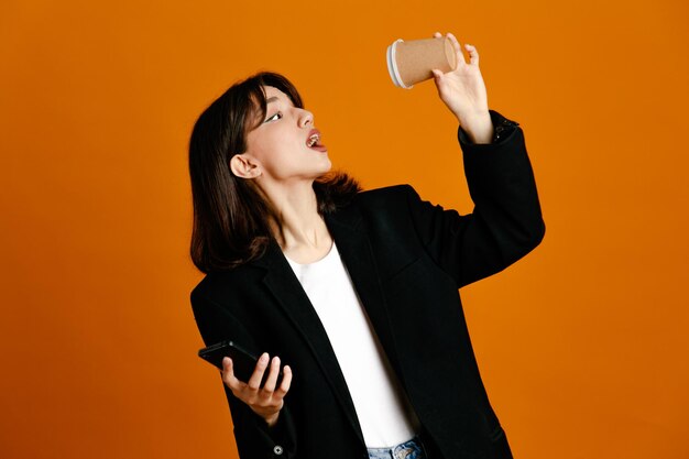 Onder de indruk met koffiekopje en telefoon jonge mooie vrouw draagt zwarte jas geïsoleerd op oranje achtergrond