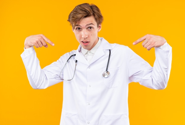 Onder de indruk van een jonge mannelijke arts die een medisch gewaad met een stethoscoop draagt, wijst naar zichzelf geïsoleerd op een oranje muur