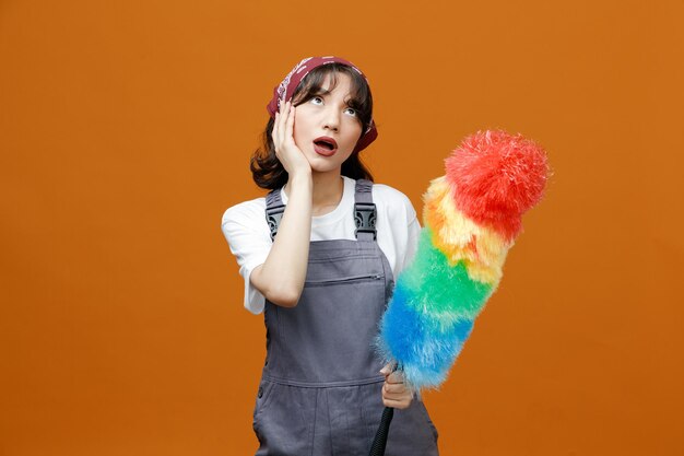 Onder de indruk van jonge vrouwelijke schoonmaker in uniform en bandana met plumeau die de hand op het gezicht houdt en omhoog kijkt geïsoleerd op een oranje achtergrond