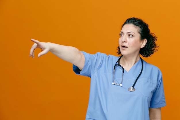 Onder de indruk vrouwelijke arts van middelbare leeftijd, gekleed in uniform en stethoscoop om haar nek, kijkend naar kant wijzend naar kant geïsoleerd op oranje achtergrond