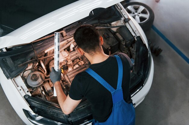 Onder de motorkap Man in werkuniform repareert witte auto binnenshuis Conceptie van autoservice