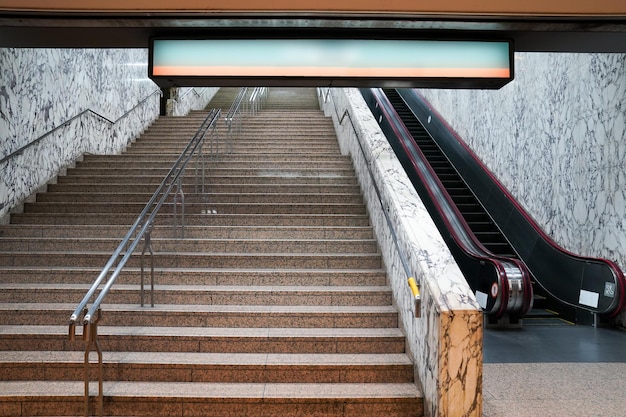 Ondergrondse poort, in- en uitgang met grote trap en roltrap ernaast. Lichtbak groen en oranje kleur is boven de poort.