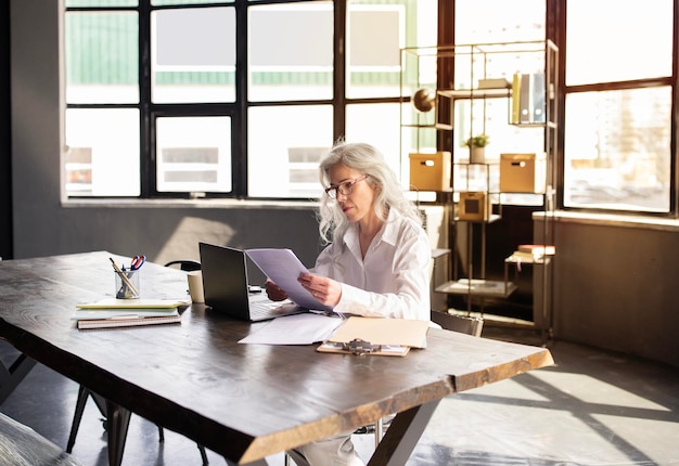 Onderneemster die bij laptop werken die handelspapieren in bureau lezen