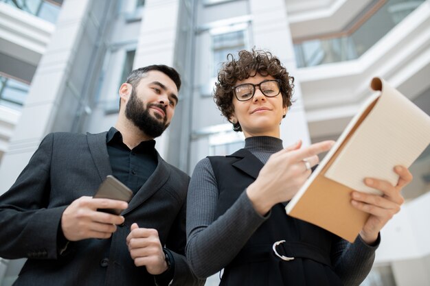 Onderneemster met krullend haar in glazen die in klembord wijzen terwijl ontwikkelingsstrategie aan collega wordt uitgelegd