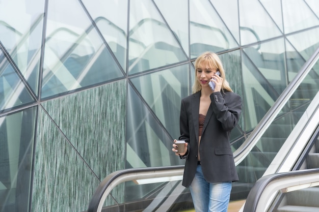 Ondernemend meisje in bedrijvenpark in een modern gebouw met roltrap, jonge blonde in een pak die belt met een afhaalkoffie