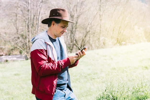 Ondernemende zelfstandige man met een hoed die naar zijn mobiele telefoon kijkt terwijl hij ontspant in het veld op een weide concept van werk geniet van ontspannen internettechnologie en connectiviteit