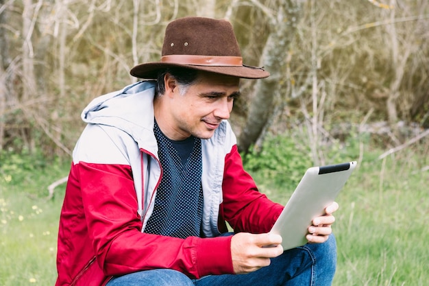 Ondernemende zelfstandige man met hoed kijkend naar zijn tablet terwijl hij ontspant in het veld zittend op een steen Concept van werk geniet van ontspannen internettechnologie en connectiviteit