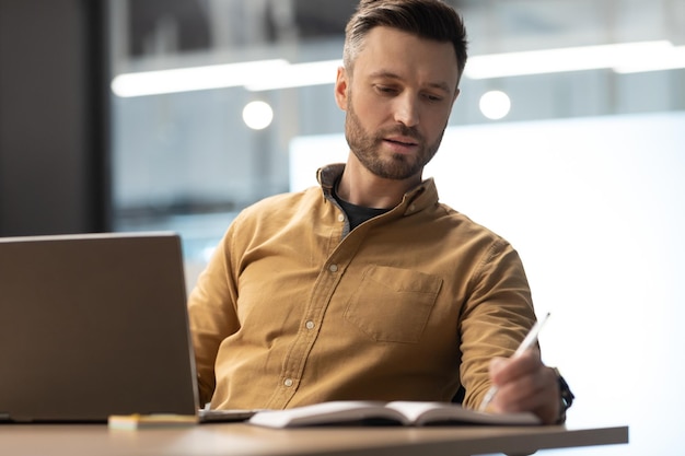 Ondernemer die online op laptop werkt en aantekeningen maakt terwijl hij op de werkplek zit