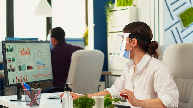 Ondernemers die tijdens het coronavirus beschermende gezichtsmaskers dragen in de kantoorruimte. Team in een nieuw normaal zakelijk financieel bedrijf dat op pc typt, rapporten controleert, gegevens analyseert en naar desktop kijkt