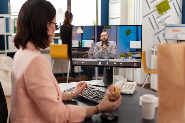 Ondernemersvrouw die aan het bureau in het kantoor van het bedrijf zit en een broodje eet tijdens een online videogesprekconferentie over financiële strategie. Afhaalbestelling eten bezorgen op zakelijke werkplek