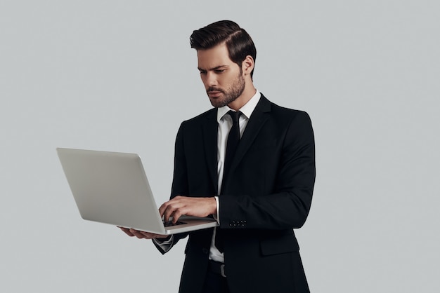 Onderweg werken. Knappe jonge man in volledig pak met laptop terwijl hij tegen een grijze achtergrond staat