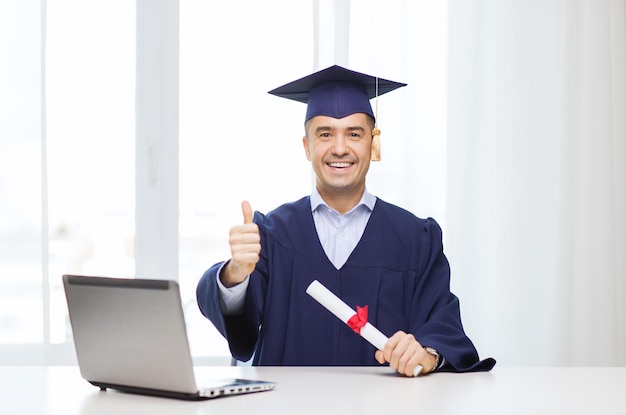 Onderwijs, afstuderen, zaken, technologie en mensen concept - gelukkige volwassen student in baret met diploma en laptop computer duimen opdagen thuis of op kantoor