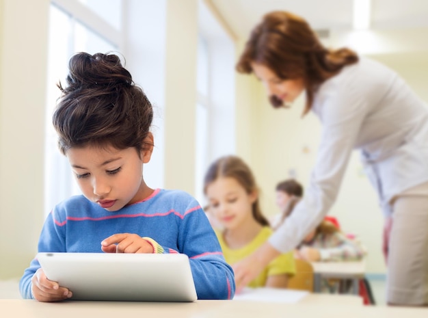 Onderwijs, basisschool, technologie en kinderenconcept - klein studentenmeisje met tabletpc over klaslokaal en leraarsachtergrond
