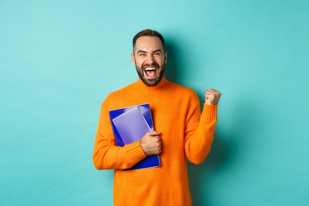 Onderwijs. Gelukkig en tevreden man verheugt zich, houdt notitieboekjes vast en maakt vuistpomp, schreeuwend van vreugde, staande tegen turkooizen achtergrond.