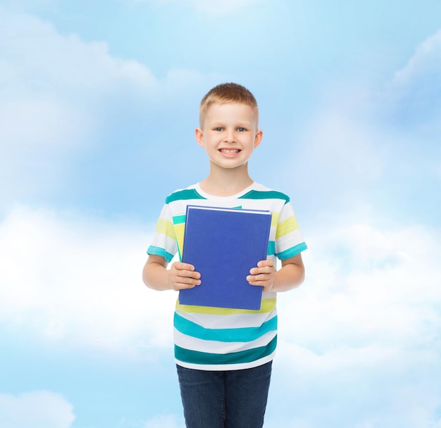 onderwijs, kindertijd en schoolconcept - glimlachende kleine studentenjongen met blauw boek over blauwe bewolkte hemelachtergrond