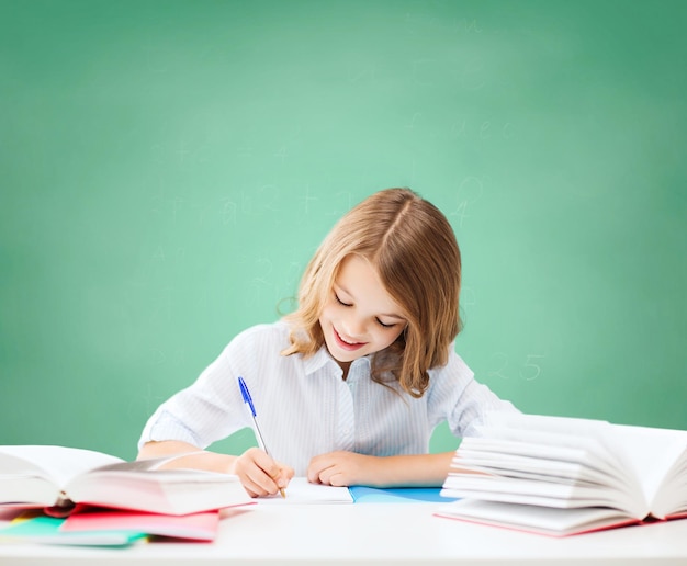 onderwijs, mensen, kinderen en school concept - gelukkig student meisje zittend aan tafel met boeken en schrijven in notitieboekje over groene krijtbord achtergrond