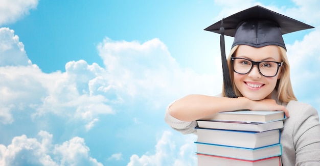 Onderwijs, middelbare school, kennis en mensen concept - foto van gelukkig student meisje of vrouw in trencher cap met stapel boeken over blauwe lucht en wolken achtergrond