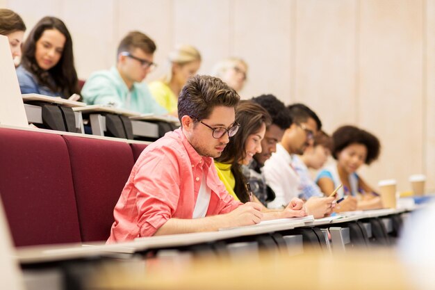 onderwijs, middelbare school, universiteit, leren en mensenconcept - groep internationale studenten die met notitieboekjes in collegezaal schrijven