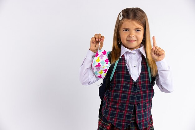 Onderwijs tijdens quarantainecovid. Kindmeisje in uniform wijst vinger naar de zijkant, geïsoleerd op grijze muur.